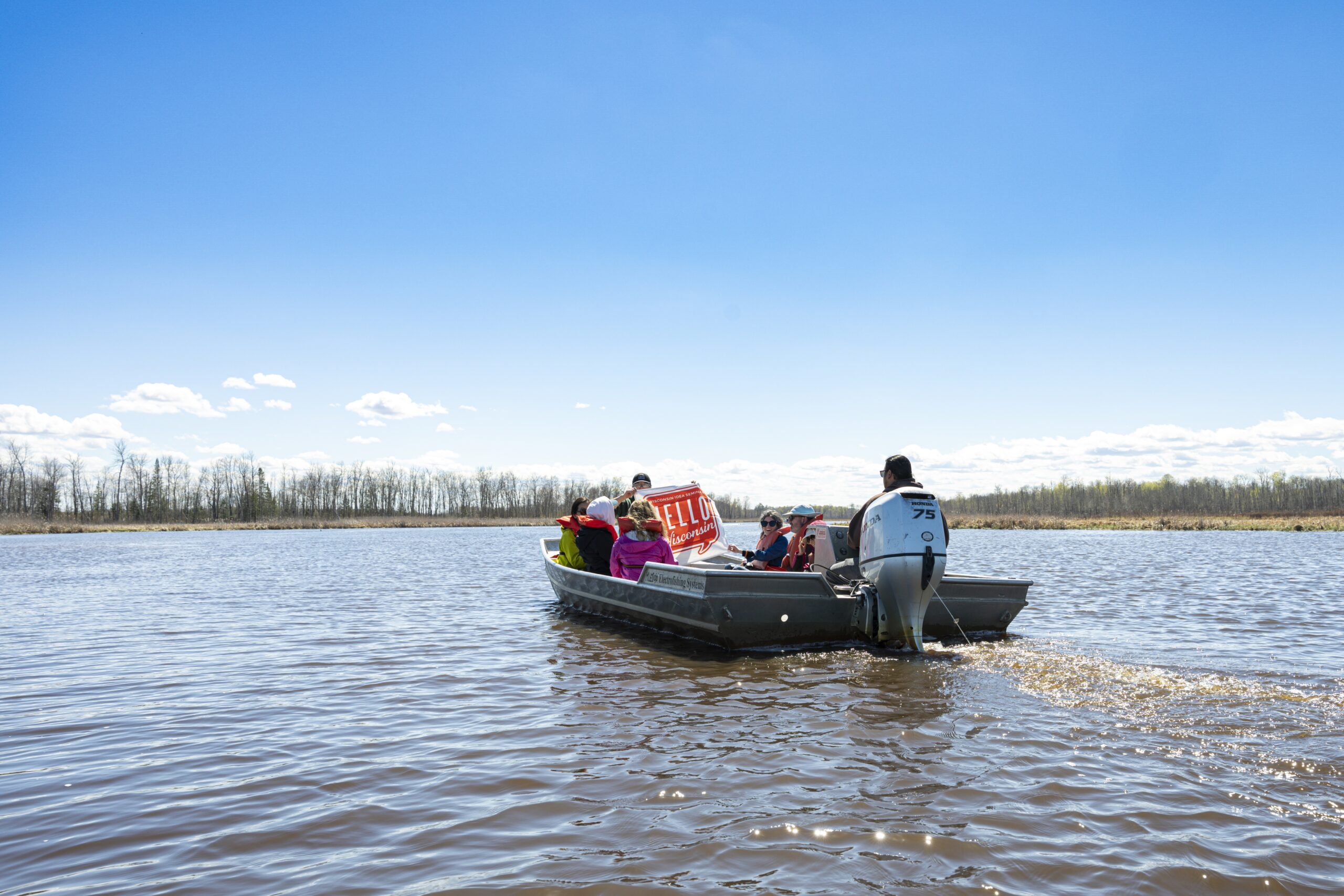Mike Wiggins Jr., tribal member of the Bad River Band of Lake Superior Chippewa, holds the Wisconsin Idea Seminar’s “Hello Wisconsin” banner as he leads a boat of 2026 cohort members over the tranquil waters of the Kakagon–Bad River Sloughs.