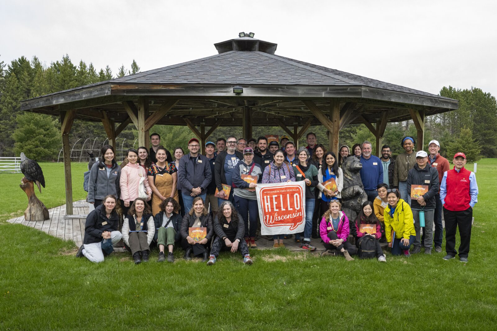 The 2026 Wisconsin Idea Seminar cohort gathers for a group photo under a gazebo on the Red Cliff Band of Lake Superior Chippewa powwow grounds. To the left, the frame of a wigwam, a round lodge, stands against the treeline.